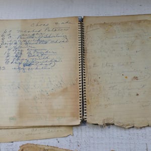 May include: An open, vintage spiral-bound notebook with handwritten recipes. The pages are aged and worn, with visible stains and tears. The left page features a list of ingredients, including measurements. The notebook is on a white wooden surface.
