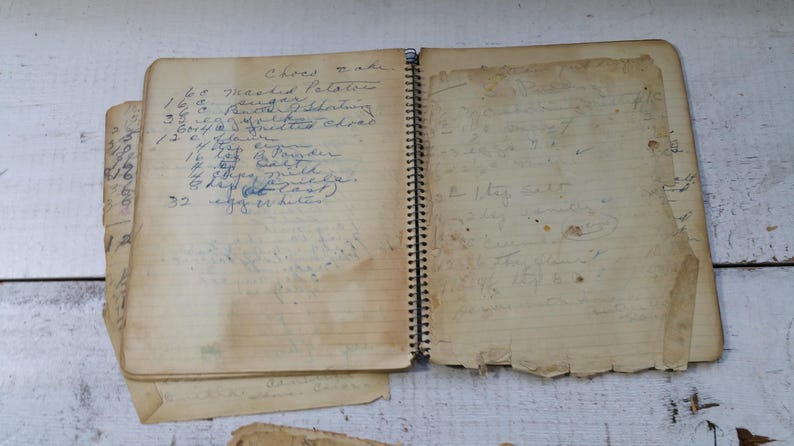 May include: An open, vintage spiral-bound notebook with handwritten recipes. The pages are aged and worn, with visible stains and tears. The left page features a list of ingredients, including measurements. The notebook is on a white wooden surface.