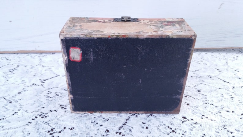 May include: A vintage wooden box with a black interior and a floral design on the top and bottom. The box has a metal latch and a small red label on the front.
