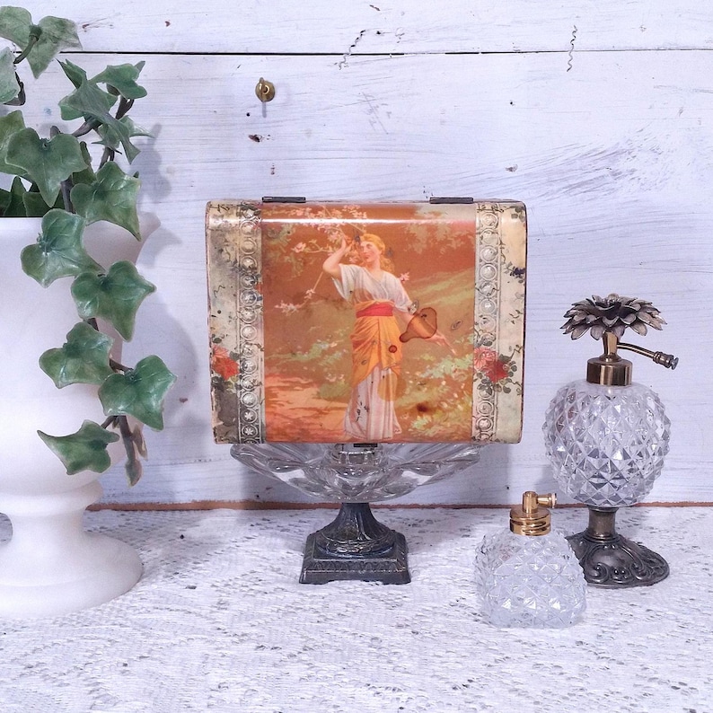 May include: A vintage tin box with a floral design and a woman in a yellow dress holding a watering can. The box is on a decorative stand with two crystal atomizers.