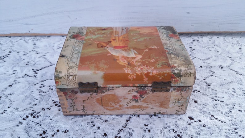 May include: A vintage wooden box with a floral design and a hinged lid. The lid features a colorful illustration of a woman holding a pitcher.