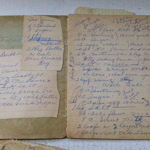 May include: An open vintage cookbook with handwritten recipes in blue ink on aged paper. The pages are yellowed, and small recipe cards are tucked inside. The cookbook is open, revealing detailed instructions.