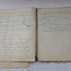 May include: A well-worn, spiral-bound notebook with handwritten recipes on aged paper. The pages are filled with faded ink, and the notebook shows signs of wear, suggesting it's been well-used. The notebook is on a white wooden surface.