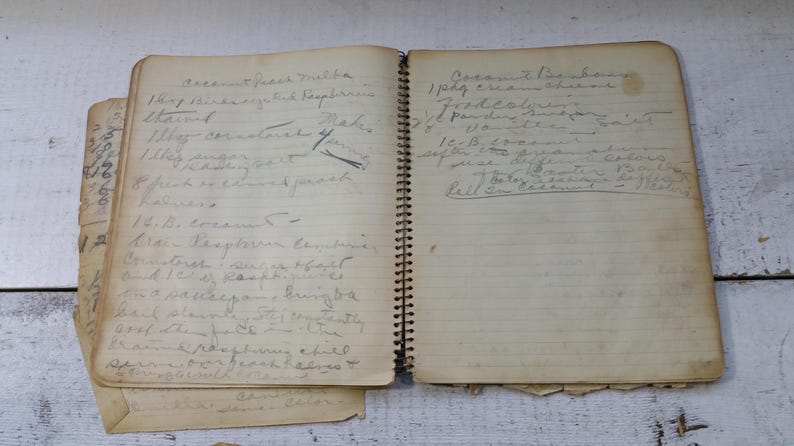 May include: A well-worn, spiral-bound notebook with handwritten recipes on aged paper. The pages are filled with faded ink, and the notebook shows signs of wear, suggesting it's been well-used. The notebook is on a white wooden surface.