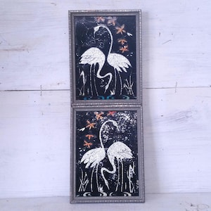 May include: Two framed vintage art pieces featuring white swans against a black background. The swans are surrounded by stylized plants and orange and red floral accents. The frames are silver.