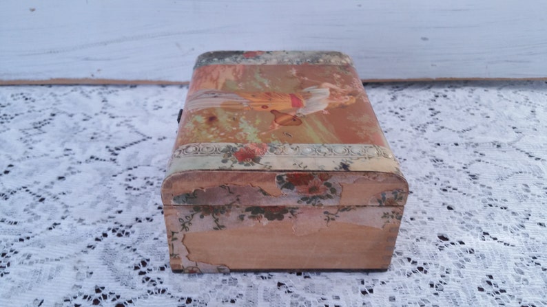 May include: A vintage wooden box with a floral design and a faded image of a woman on the lid. The box is in a worn condition, with some of the paper peeling off.