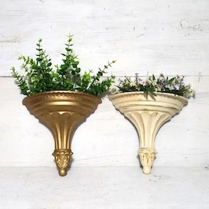 May include: Two decorative wall sconces, one gold and one cream, each holding greenery and flowers. The sconces have a classic, ornate design with a scalloped top and a detailed base, perfect for displaying plants or small decor items.