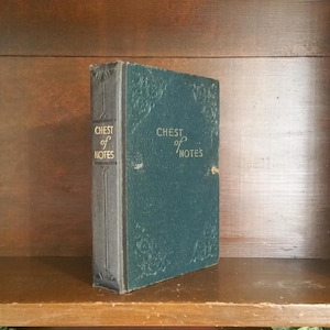 Puede incluir: Un diario vintage de color verde azulado titulado "Chest of Notes". El libro tiene detalles en relieve ornamentados en la portada y el lomo. El lomo es de un color gris más oscuro. El diario está de pie sobre una estantería de madera.