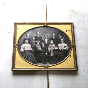May include: A vintage oval-shaped photograph of a family portrait in a gold frame. The photo shows a group of 10 people, including adults and children, dressed in formal attire. The photo is in black and white and has a faded look.