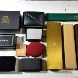 May include: A collection of vintage watch and jewelry boxes in various shapes and colors. Boxes include black, gold, green, and red velvet. Some boxes have brand names like Bulova, Speidel, and Waltham.