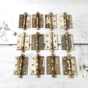 以下が含まれることがあります： 12個のヴィンテージメタルドアヒンジのセット。各ヒンジは長方形のベースと中央のピンを備えています。白とグレーの風合いのある塗装が施されており、使用感が見られます。素朴またはアンティークなスタイルを連想させます。