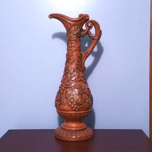 May include: A tall, brown decorative pitcher with a handle and spout. The pitcher is adorned with grape and leaf relief designs, and gold accents. The base is round and detailed. The pitcher is approximately 38 cm tall.