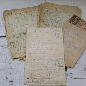 May include: A collection of aged, handwritten recipes and notes on aged paper. The papers are arranged on a white, distressed wooden surface. Some papers have visible text, including a recipe for "9 bread hammon" and a "Still's" tuna and chicken advertisement.