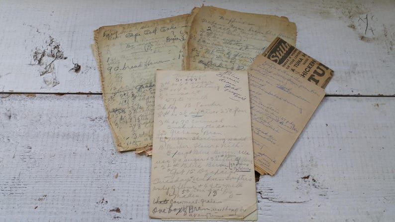 May include: A collection of aged, handwritten recipes and notes on aged paper. The papers are arranged on a white, distressed wooden surface. Some papers have visible text, including a recipe for "9 bread hammon" and a "Still's" tuna and chicken advertisement.