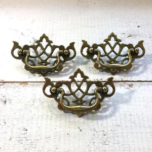 Könnte beinhalten: Drei antike Schubladengriffe aus Messing mit aufwendigen, durchbrochenen Designs. Jeder Griff verfügt über einen gebogenen Griff und dekorative Details, ideal, um Möbeln einen Vintage-Touch zu verleihen. Die Griffe sind auf einer weißen Holzoberfläche angeordnet.