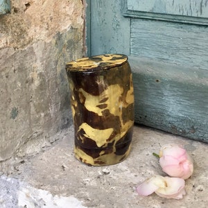 Pode incluir: Um recipiente cilíndrico de cerâmica com tampa, com um padrão marmorizado castanho e amarelo. O recipiente está sobre uma superfície de concreto ao lado de uma porta azul clara e uma parede desgastada. Duas pétalas de rosa rosa estão por perto.