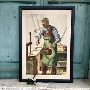 May include: Framed print showing an elderly artisan in a workshop, wearing a green apron and working at a wooden table. The print is in a black frame and rests on a stone step. A small bouquet of white flowers is placed in front of the frame.