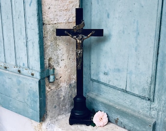 May include: A black crucifix with a bronze Jesus figure stands on a stone ledge. The cross is positioned in front of a weathered blue door and a light pink rose rests nearby. The cross has a decorative gold band.