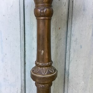 May include: A close-up of a bronze-colored, ornate metal stand. The stand features decorative floral and circular patterns along its column. The top portion is a bowl shape. The stand has a weathered appearance.