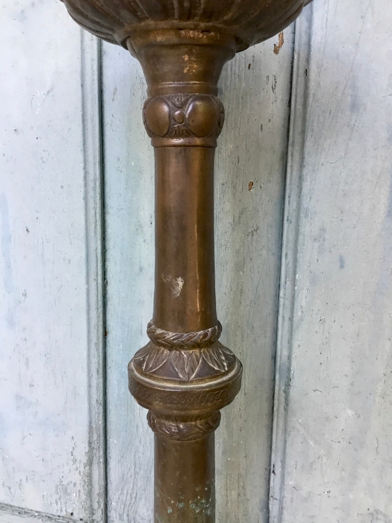 May include: A close-up of a bronze-colored, ornate metal stand. The stand features decorative floral and circular patterns along its column. The top portion is a bowl shape. The stand has a weathered appearance.