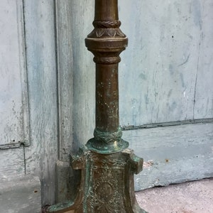 May include: An antique bronze candlestick with a detailed, ornate base and a tall, cylindrical stem. The candlestick has a weathered patina, with hints of verdigris, suggesting age and exposure to the elements. A pink rose lies at the base.