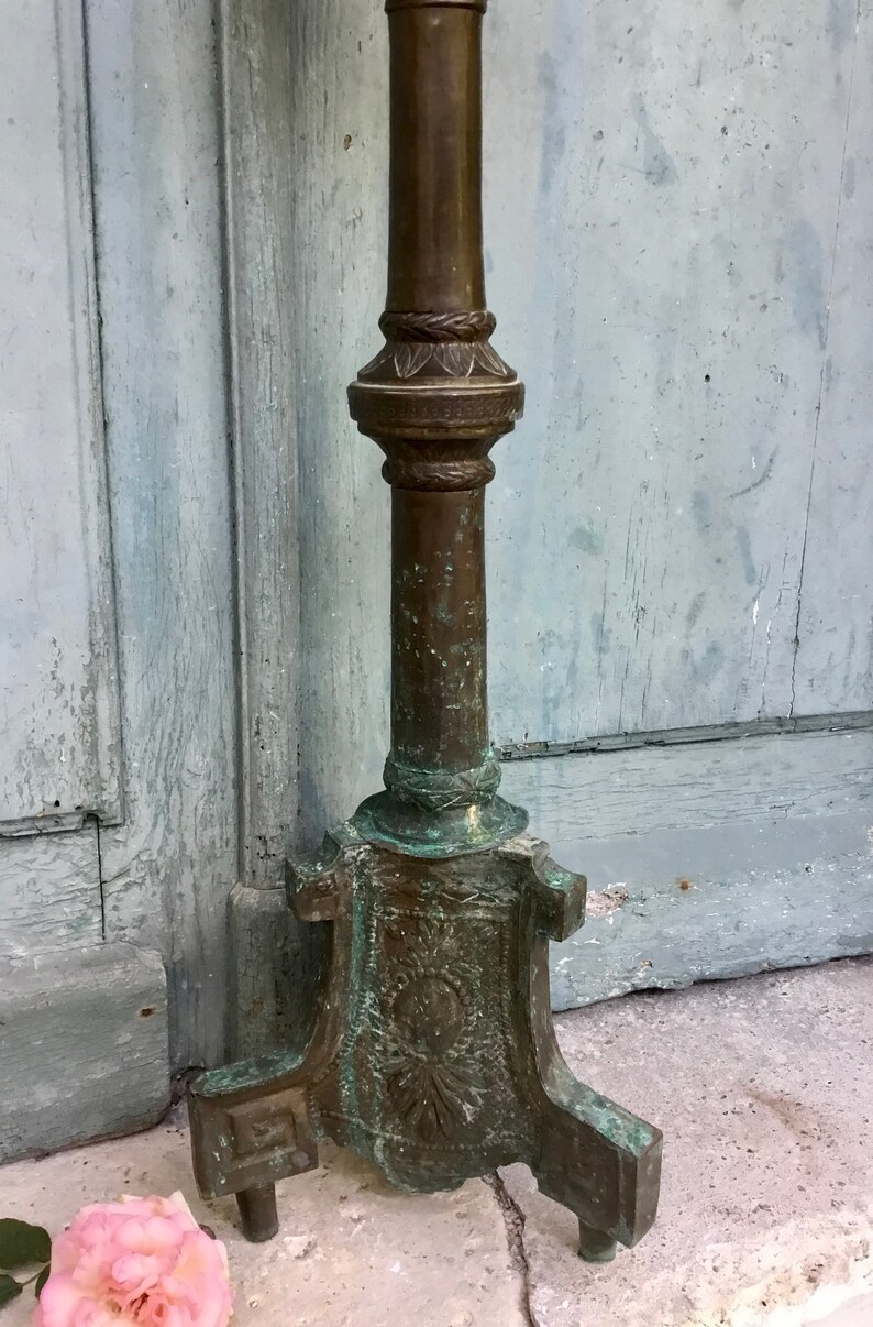 May include: An antique bronze candlestick with a detailed, ornate base and a tall, cylindrical stem. The candlestick has a weathered patina, with hints of verdigris, suggesting age and exposure to the elements. A pink rose lies at the base.