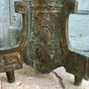 May include: Close-up of a bronze-colored decorative stand base with ornate floral and geometric designs. The stand has a weathered, aged appearance with hints of green patina. The base features three short legs and a detailed central panel.