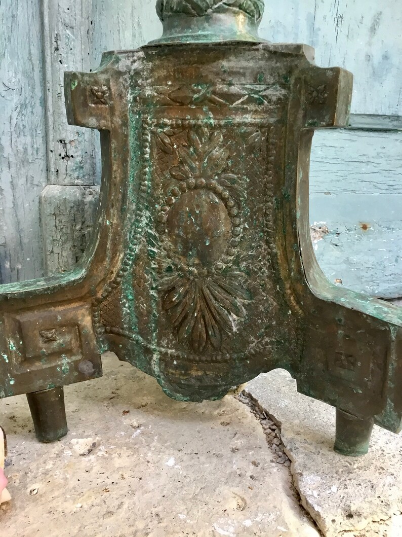May include: Close-up of a bronze-colored decorative stand base with ornate floral and geometric designs. The stand has a weathered, aged appearance with hints of green patina. The base features three short legs and a detailed central panel.