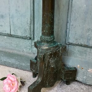 May include: An antique bronze-colored metal stand with a decorative column and base. The stand has a weathered appearance with green patina. A pink rose lies on the ground near the stand.