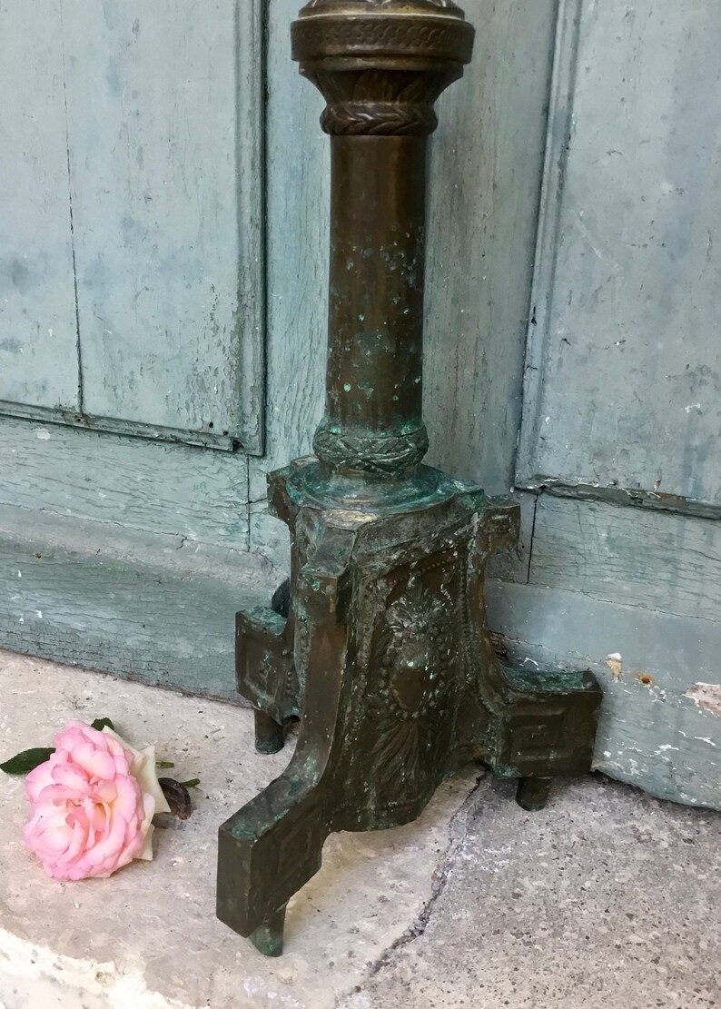 May include: An antique bronze-colored metal stand with a decorative column and base. The stand has a weathered appearance with green patina. A pink rose lies on the ground near the stand.
