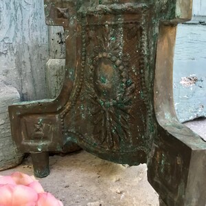 May include: Close-up of a weathered bronze decorative object with ornate floral and geometric relief details. The object has a patina of green and brown hues, with three short legs. A pink rose is in the foreground.