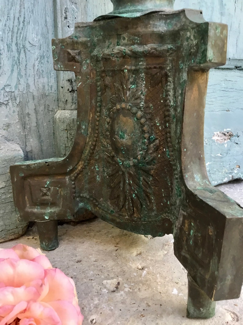 May include: Close-up of a weathered bronze decorative object with ornate floral and geometric relief details. The object has a patina of green and brown hues, with three short legs. A pink rose is in the foreground.