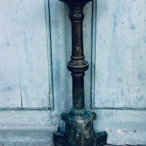 May include: An antique bronze lamp with a detailed, ornate design. The lamp has a tall, slender stem and a wide, shallow bowl at the top. The base is a tripod design. A pink rose lies on the ground near the lamp.