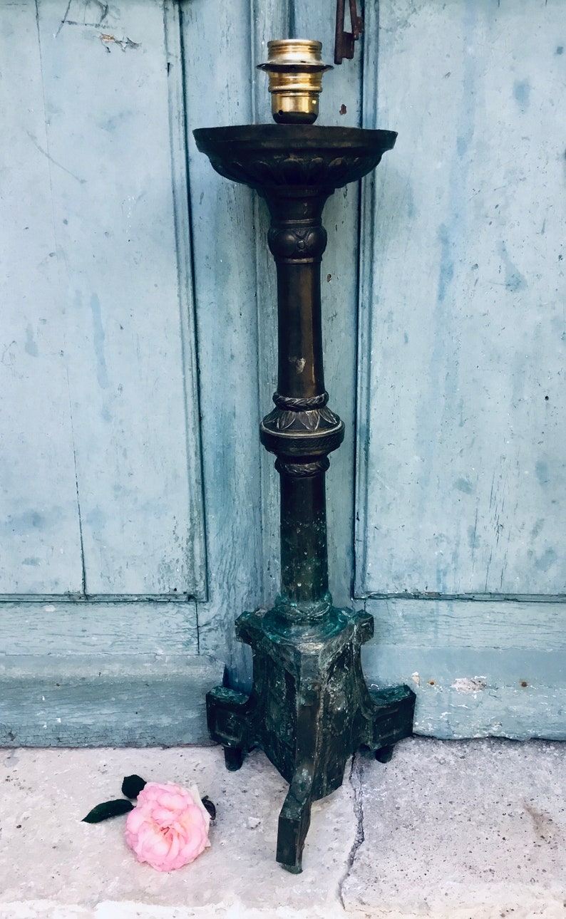 May include: An antique bronze lamp with a detailed, ornate design. The lamp has a tall, slender stem and a wide, shallow bowl at the top. The base is a tripod design. A pink rose lies on the ground near the lamp.