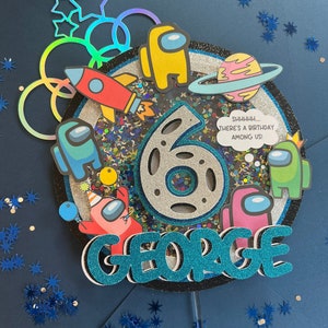 May include: A blue and silver cake topper with a silver number 6, the words "GEORGE" and "SHHHH... THERE'S A BIRTHDAY AMONG US!" in a speech bubble. The topper is decorated with colorful glitter, stars, and characters from the video game Among Us.