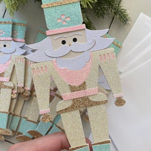 May include: A set of nutcracker ornaments made of glitter paper. The nutcrackers are in shades of pink, white, gold, and teal. The nutcrackers are standing upright and have a festive, whimsical look.