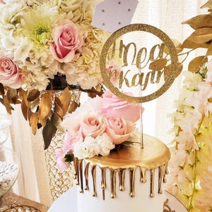 May include: A white cake with gold drip icing and a gold glitter ring topper that says "Mea & Kayden". The cake is decorated with pink and white flowers.