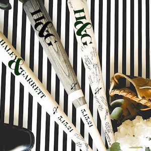 May include: Three white baseball bats with black and green lettering. The bats are decorated with the names "Haley & Garrett" and "H&G" and the date "5.15.21". The bats are lying on a black and white striped surface.