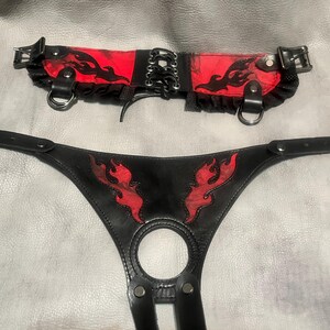 May include: Black leather harness and collar set with red flame accents. The collar features a red fabric panel with black flame designs, lace-up detailing, and metal hardware. The harness has a circular opening and matching flame designs.
