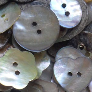 May include: A collection of iridescent mother-of-pearl buttons in various shapes and sizes, including round, flower, and heart shapes. The buttons have a subtle shimmer and a pearly sheen.