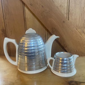 May include: A vintage tea set comprising a silver-toned teapot and creamer with white detailing. The teapot has a curved handle and a decorative lid. The creamer has a small handle and spout. The set is displayed on a wooden surface.