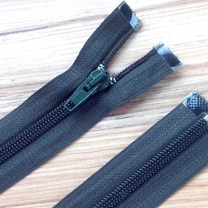 May include: Two black zipper closures with a green slider on one and a silver slider on the other. The zippers are laid out on a wooden surface.