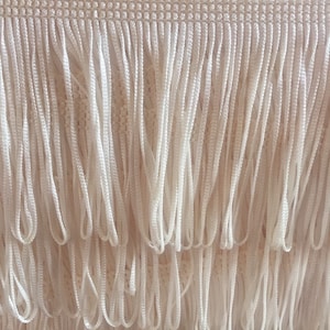 May include: White fringe trim with a woven top edge. The fringe is made of thin, white cords.