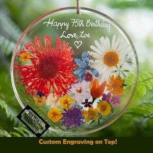 Peut inclure: Ornement circulaire transparent avec des fleurs pressées, dont une grande marguerite rouge. L'ornement porte l'inscription "Happy 75th Birthday Love, Zoe" et un cœur. Une petite étiquette indique "Natural Flowers".