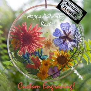 May include: A clear, circular ornament filled with colorful dried flowers, including red, blue, yellow, and purple blooms. The ornament has the text "Happy Mother's Day! Rachel" and "Custom Engraving!" with a "Natural Flowers" label.