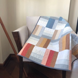 May include: A patchwork quilt with a geometric pattern in shades of blue, brown, beige, and pink. The quilt is made of linen fabric and has a simple, modern design.