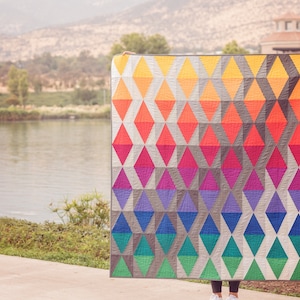 Op de afbeelding: Een kleurrijk quilt met een geometrisch patroon van ruiten in tinten geel, oranje, rood, roze, paars, blauw en groen. De quilt is gemaakt met een witte achtergrond en grijze accenten.