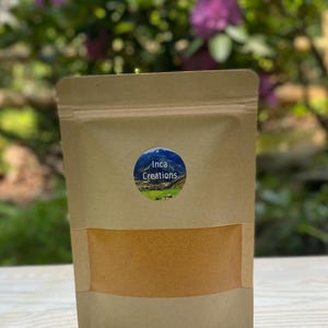 May include: A brown paper pouch with a clear window, filled with a light orange substance. The pouch has a blue and white circular sticker with the text "Inca Creations". The background features a wooden table and green foliage.