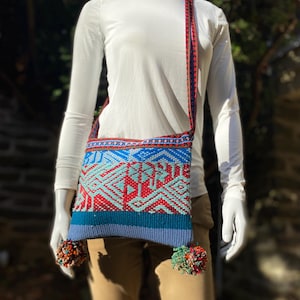 May include: A woven shoulder bag with a geometric pattern in red, blue, and turquoise. The bag has a long strap with a red and blue design and is adorned with two pom-poms at the bottom. The bag is displayed on a mannequin.