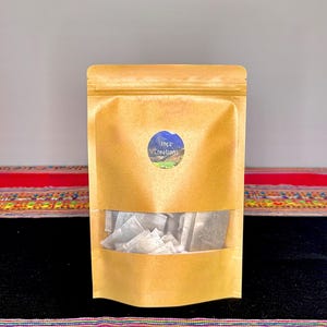 May include: A brown paper bag with a clear window, filled with tea bags. The bag has a blue and white circular sticker that reads "Inca Creations". The bag is sitting on a colorful textile.